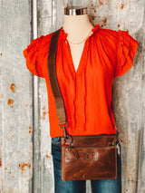 Photo of terra cotta red top with short ruffled sleeve and a v neck on a mannequin styled with a brown leather crossbody purse with a tin background