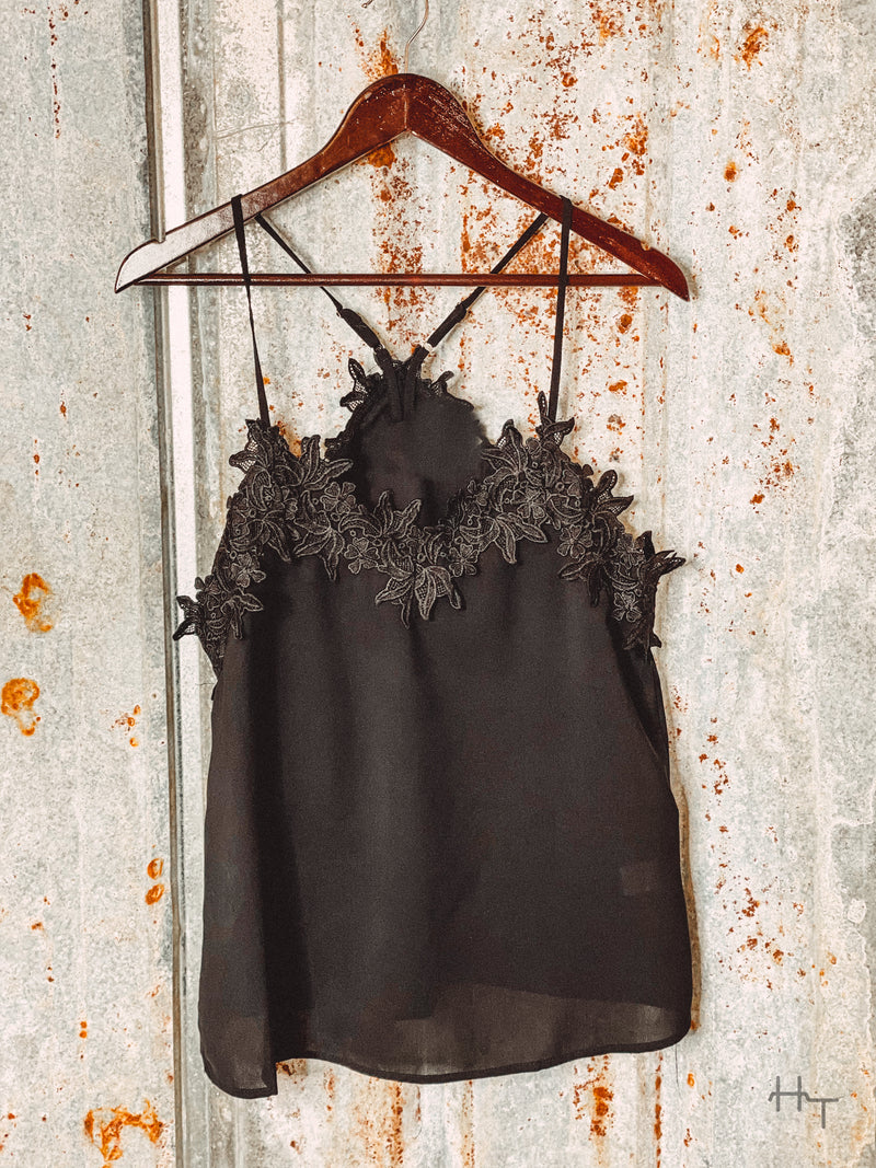 Photo of front view of black cami with floral lace detail on hanger with tin background