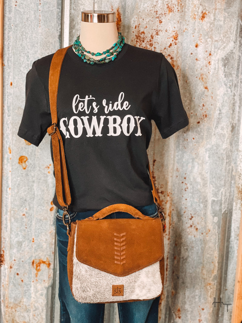 Photo of black tee shirt with Let's Ride Cowboy on front in white paired with turquoise necklace and leather and white cowhide purse on a mannequin with tin background