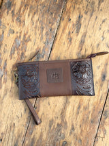 Photo of front of dark brown leather clutch wallet with tooled leather on the sides and STS patch on front  with wood back ground