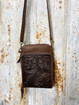 Photo of close up of front of chocolate brown leather purse with tooled leather panel on front pocket with long crossbody strap with tin background
