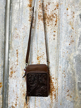 Photo of front of chocolate brown leather purse with tooled leather panel on front pocket with long crossbody strap with tin background