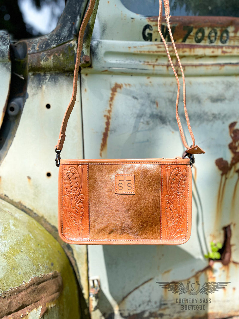 Image of the front of the  Yipee Kiyay Claire crossbody purse with crossbody strap.  The purse has a brown cowhide panel in the middle with floral tooled veggie tan leather on each side and a leather patch in the middle with the STS brand.