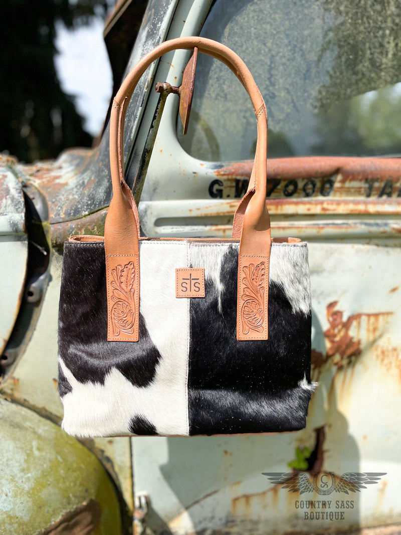 Image of yipee kiyay tote purse with black and white cowhide front with veggie tan leather rolled shoulder straps with tooled leather detail. 